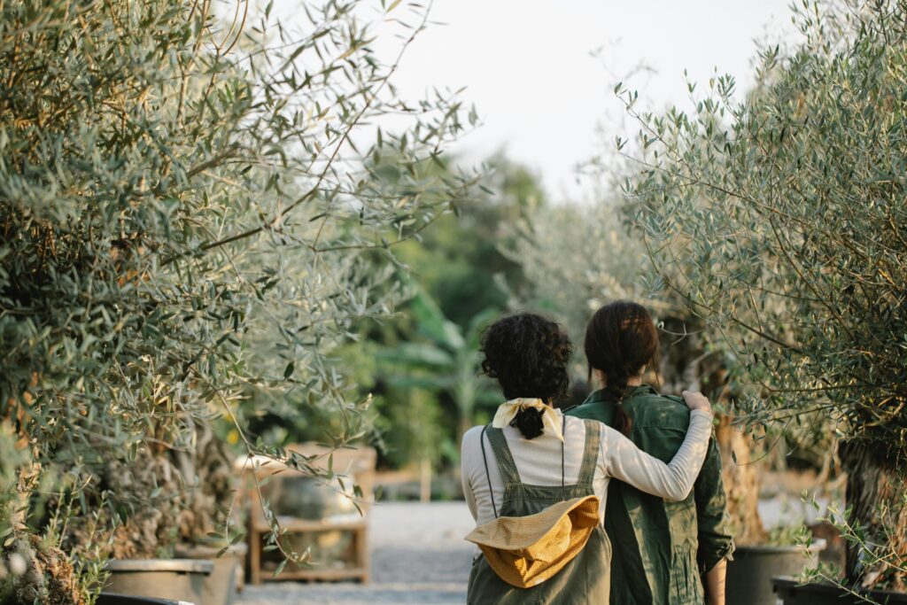 Back view of anonymous female gardeners hugging while standing in orangery with various green plants on blurred background during work